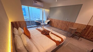 A modern bedroom with a large bed, wooden paneling, a small table, and a view of the city through large windows. - Ikonic Hotel - Bhushi Dam Lonavala (Lonavala)