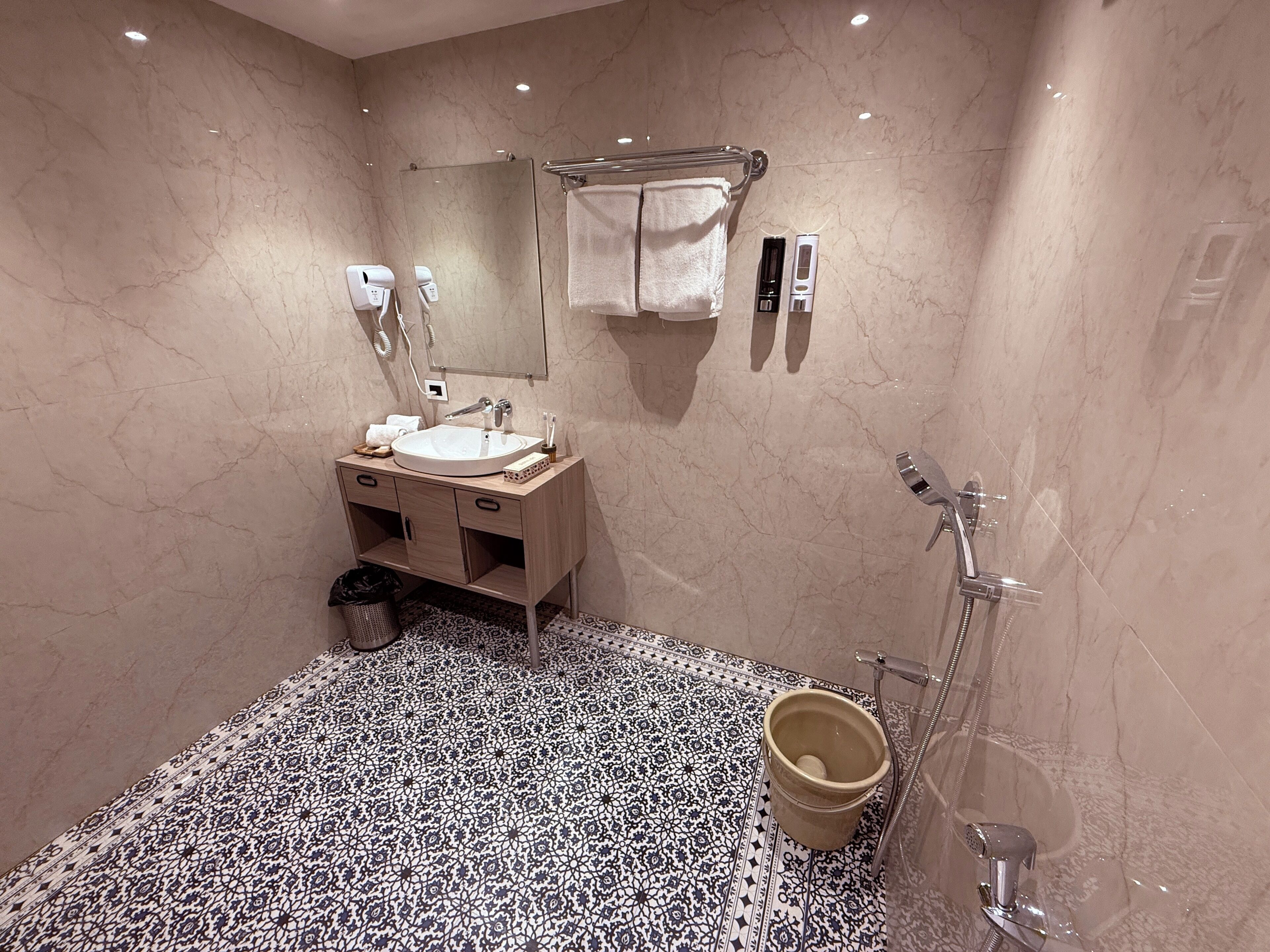 A modern bathroom with a marble vanity, a large mirror, a towel rack, a handheld showerhead, and a patterned floor.