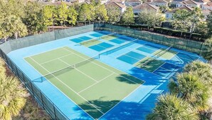 A tennis court with multiple courts, surrounded by a fence, and residential houses in the background.