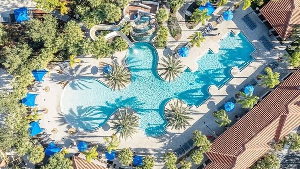 An aerial view of a resort with a central swimming pool surrounded by palm trees, lounge chairs, and a water slide.