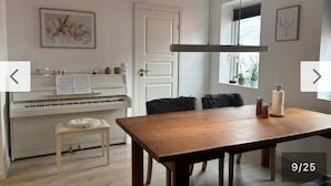 Una habitación con un piano, una mesa de comedor de madera, sillas negras, un banco mullido y una ventana con plantas.