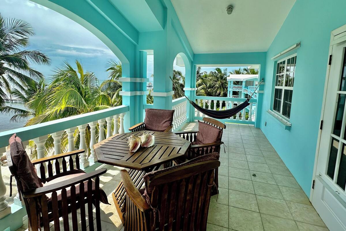 A turquoise balcony with a wooden table and chairs, a hammock, and a view of palm trees and the sea.