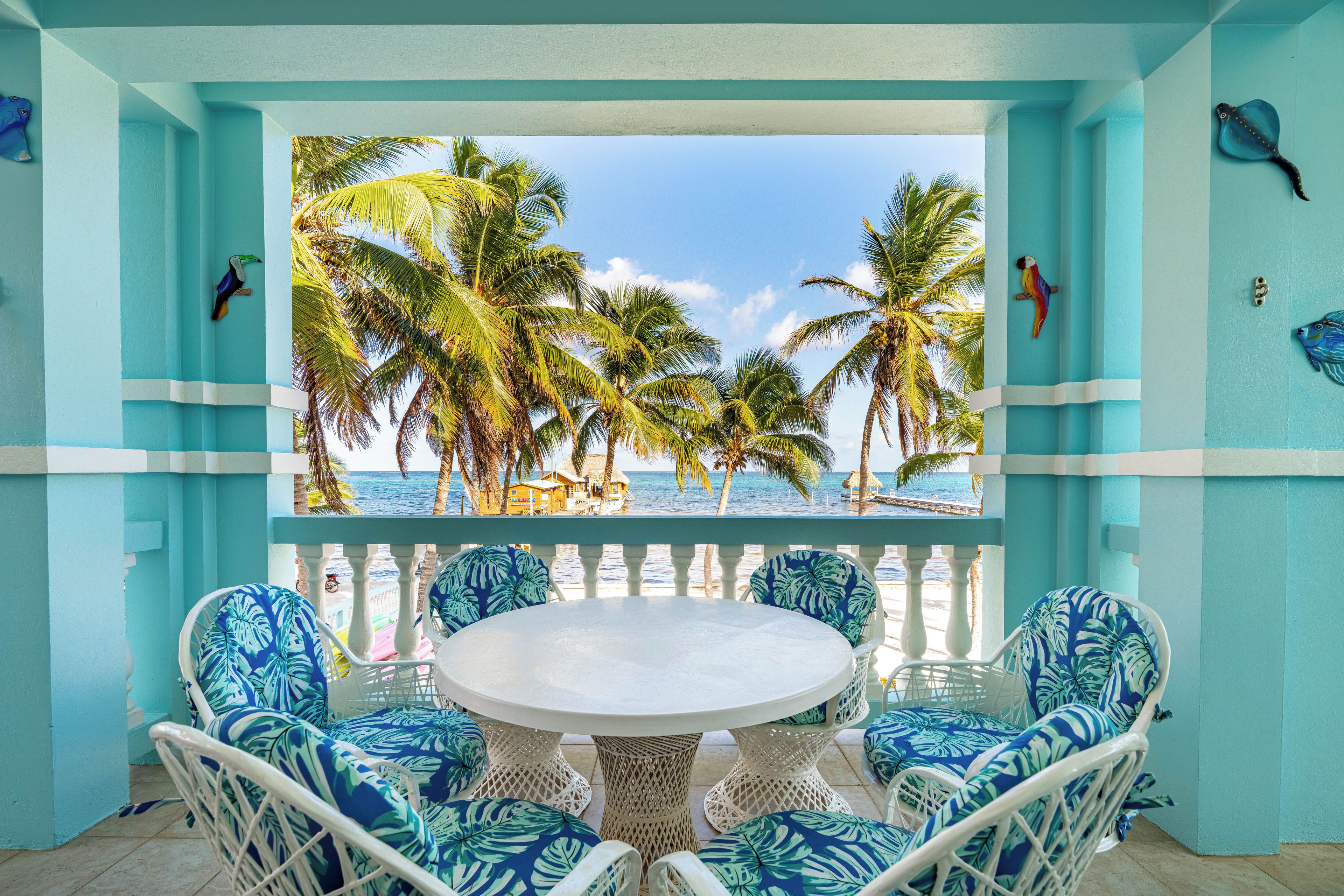 A turquoise-colored room with a round table and chairs, a view of palm trees and the sea, and a thatched-roof building in the background.