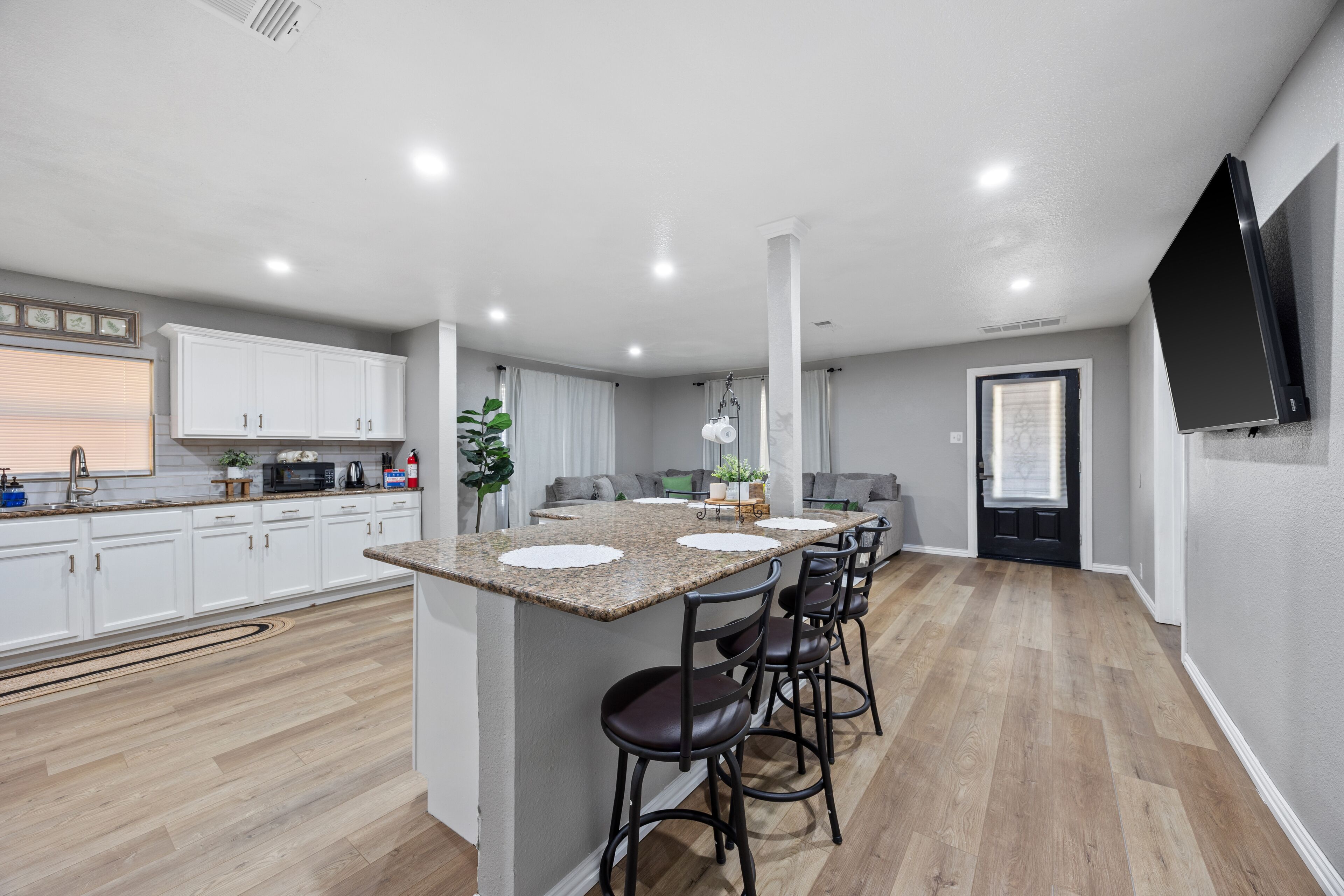 A modern kitchen with a central island, bar stools, white cabinets, and a flat-screen TV mounted on the wall.