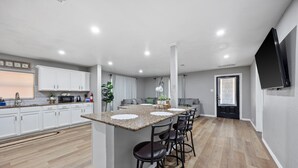 Een moderne keuken met een centraal eiland, barstoelen, witte kasten en een flatscreen televisie aan de wand.