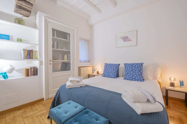 A bedroom with a large bed, a bedside table, a nightstand, a bookshelf, and a window.