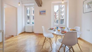 A dining area with a round table, white chairs, and a view of buildings through large windows. - Sweett | Moscova 2b - Two Bedroom Apartment, Sleeps 5 (Milano)