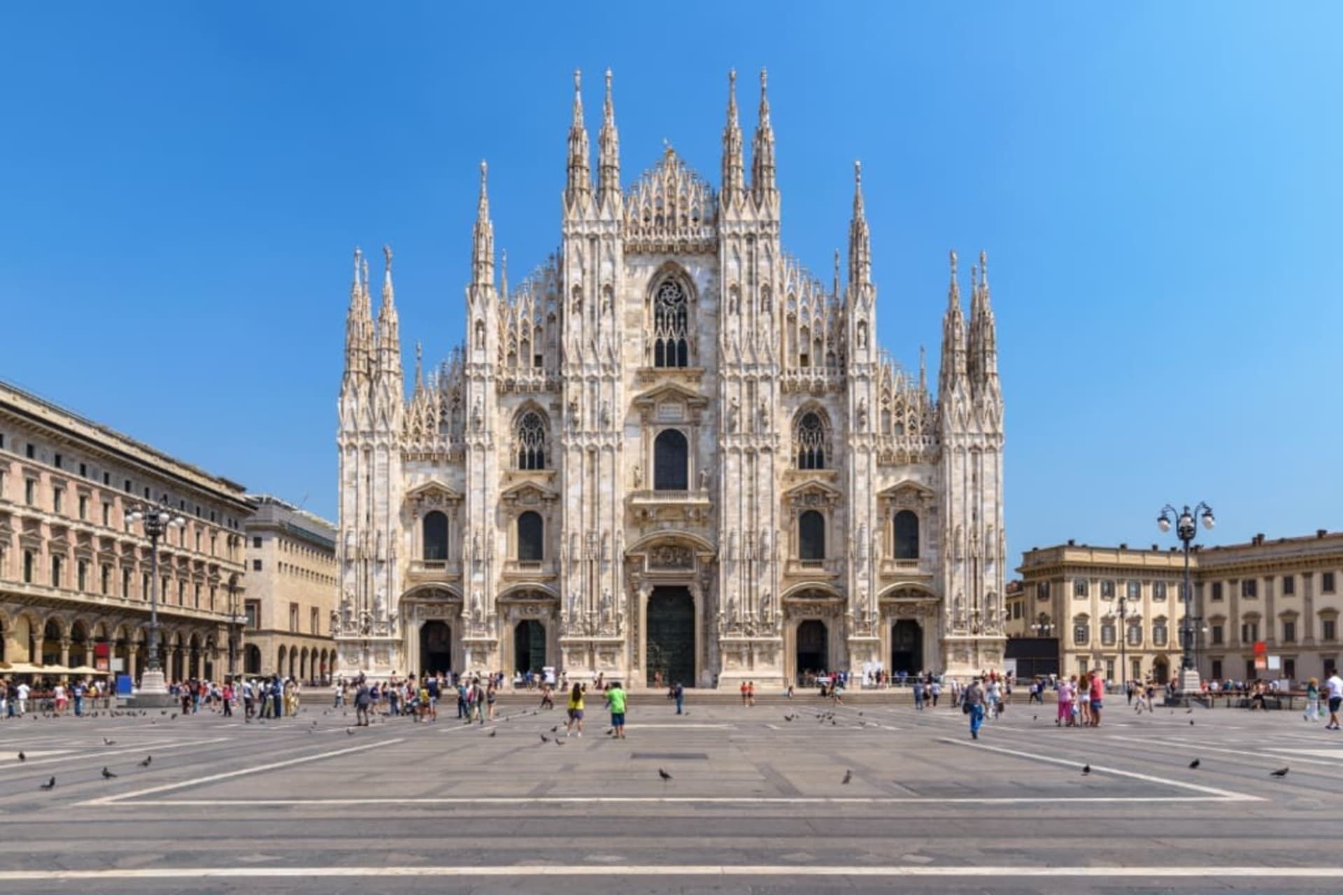 A large cathedral with a central dome and multiple spires, surrounded by a spacious plaza with people and pigeons.