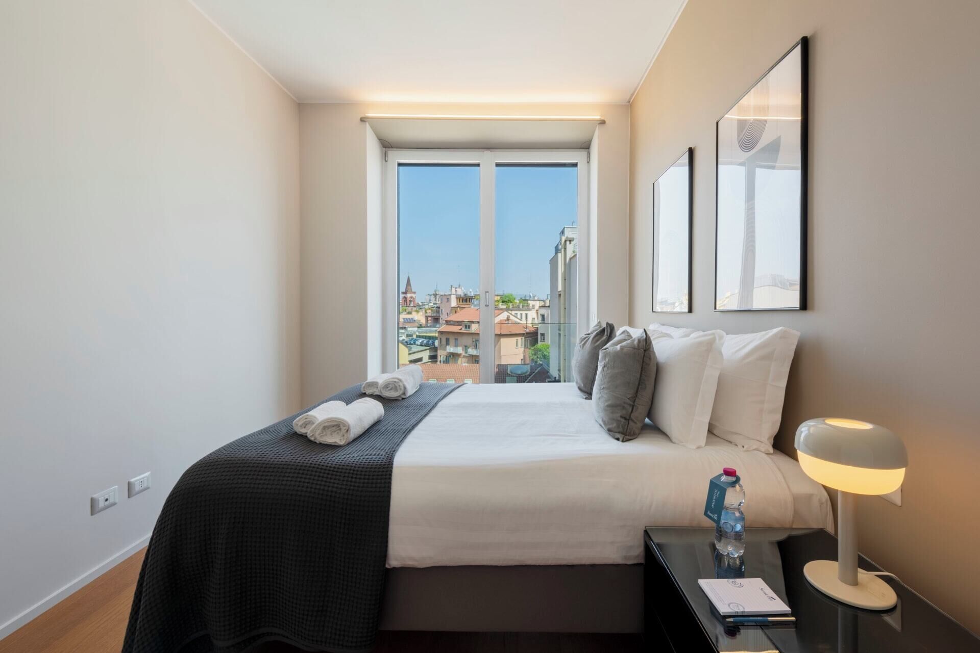 A hotel room with a bed, a bedside table with a lamp, a water bottle, and a view of the city through large windows.