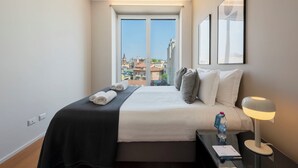 A hotel room with a bed, a bedside table with a lamp, a water bottle, and a view of the city through large windows. - Sweett | Luxury Velasca IV (Milano)