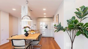 A modern kitchen with white cabinets, a built-in oven, and a dining area with a wooden table and metal chairs. - Sweett | Luxury Velasca IV (Milano)