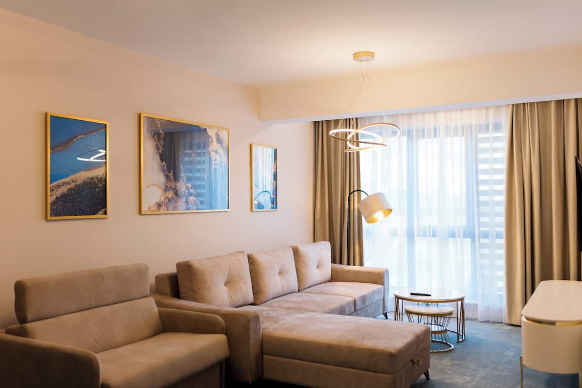 Une chambre d’hôtel moderne avec un canapé d’angle, une petite table ronde et un fauteuil. Trois œuvres d’art encadrées ornent le mur, et une grande fenêtre est agrémentée de rideaux.