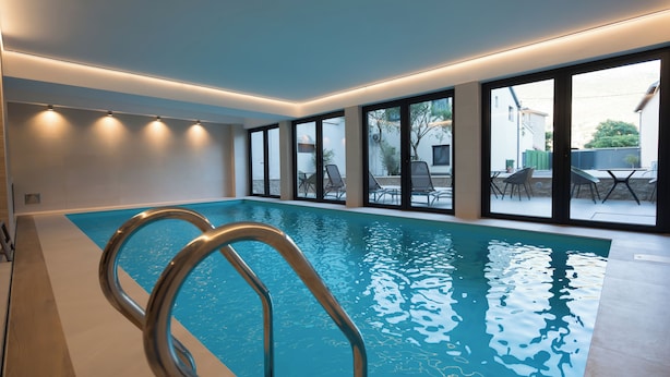 An indoor swimming pool with clear glass walls, a metal pool ladder, and a view of the outside patio area.