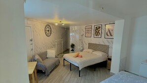 A bedroom with a bed, a desk, and a chair. There is a window with curtains, a wall clock, and framed pictures on the wall.