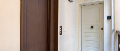 A hallway with a closed brown door, a white door with a number plate, and a light switch on the wall.