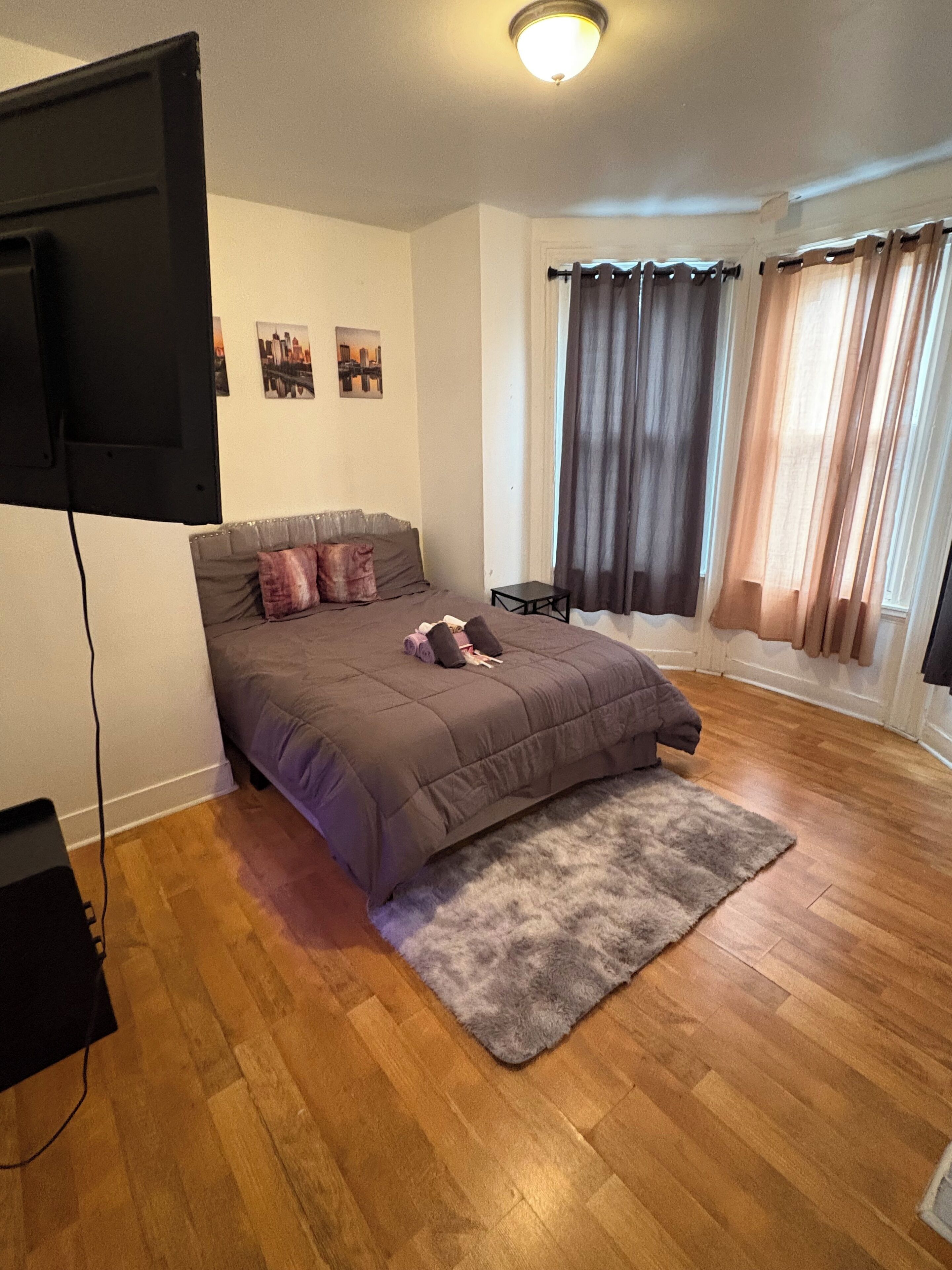 Ein Schlafzimmer mit einem Bett, einem an der Wand montierten Fernseher, einem kleinen Tisch und einem Fenster mit Vorhängen.