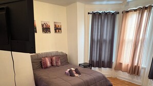 Ein Schlafzimmer mit einem Bett, einem an der Wand montierten Fernseher, einem kleinen Tisch und einem Fenster mit Vorhängen.