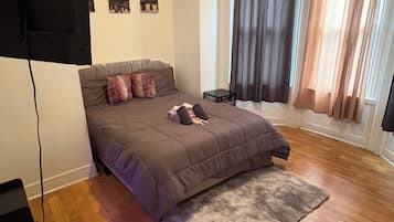 Ein Schlafzimmer mit einem Bett, einem an der Wand montierten Fernseher, einem kleinen Tisch und einem Fenster mit Vorhängen.
