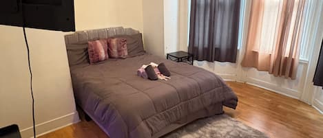 A bedroom with a bed, a TV mounted on the wall, a small table, and a window with curtains.