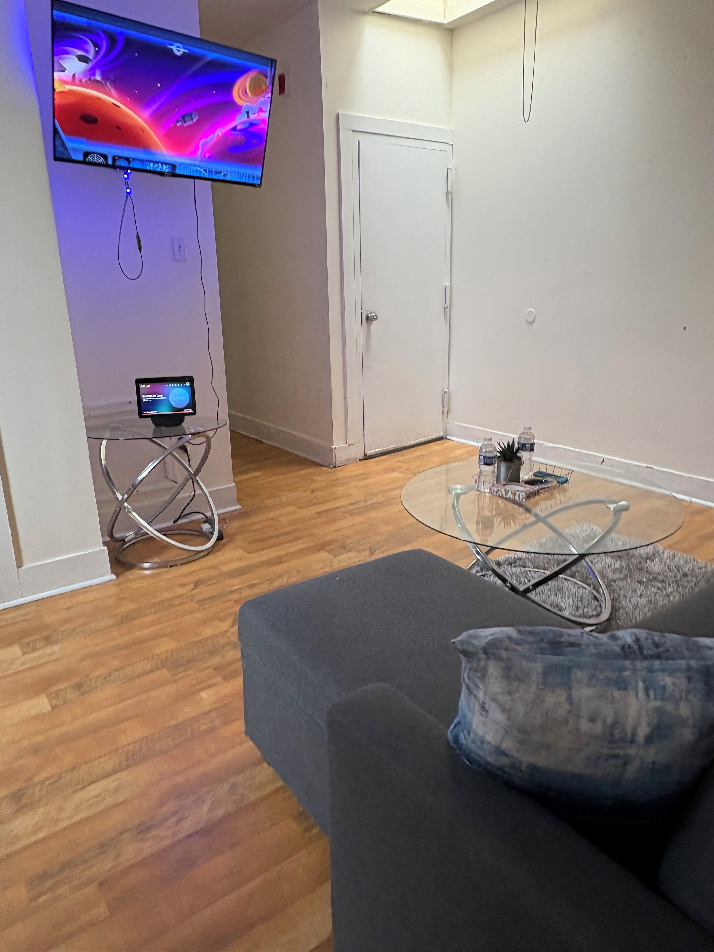 A modern living room with a flat-screen TV mounted on the wall, a glass coffee table, a dark sofa, and a small side table with a remote control.