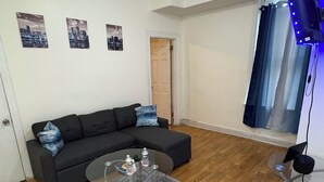 Ein Wohnzimmer mit einem dunkelgrauen Ecksofa, einem Glastisch und einem grauen Teppich. An der Wand hängen drei gerahmte Stadtansichten, der Boden ist Holzboden und ein Fenster mit blauen LED-Leuchten.