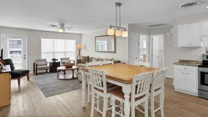 Een ruime woonkamer met een houten eettafel, witte stoelen, een bank en een salontafel. Er is een keuken met witte kastjes en een fornuis.