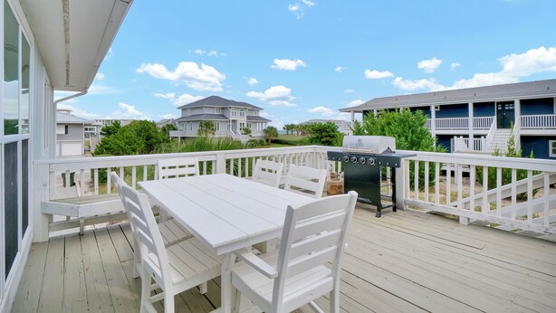 Een houten terras met een witte tafel en stoelen, een barbecue en uitzicht op omliggende huizen.