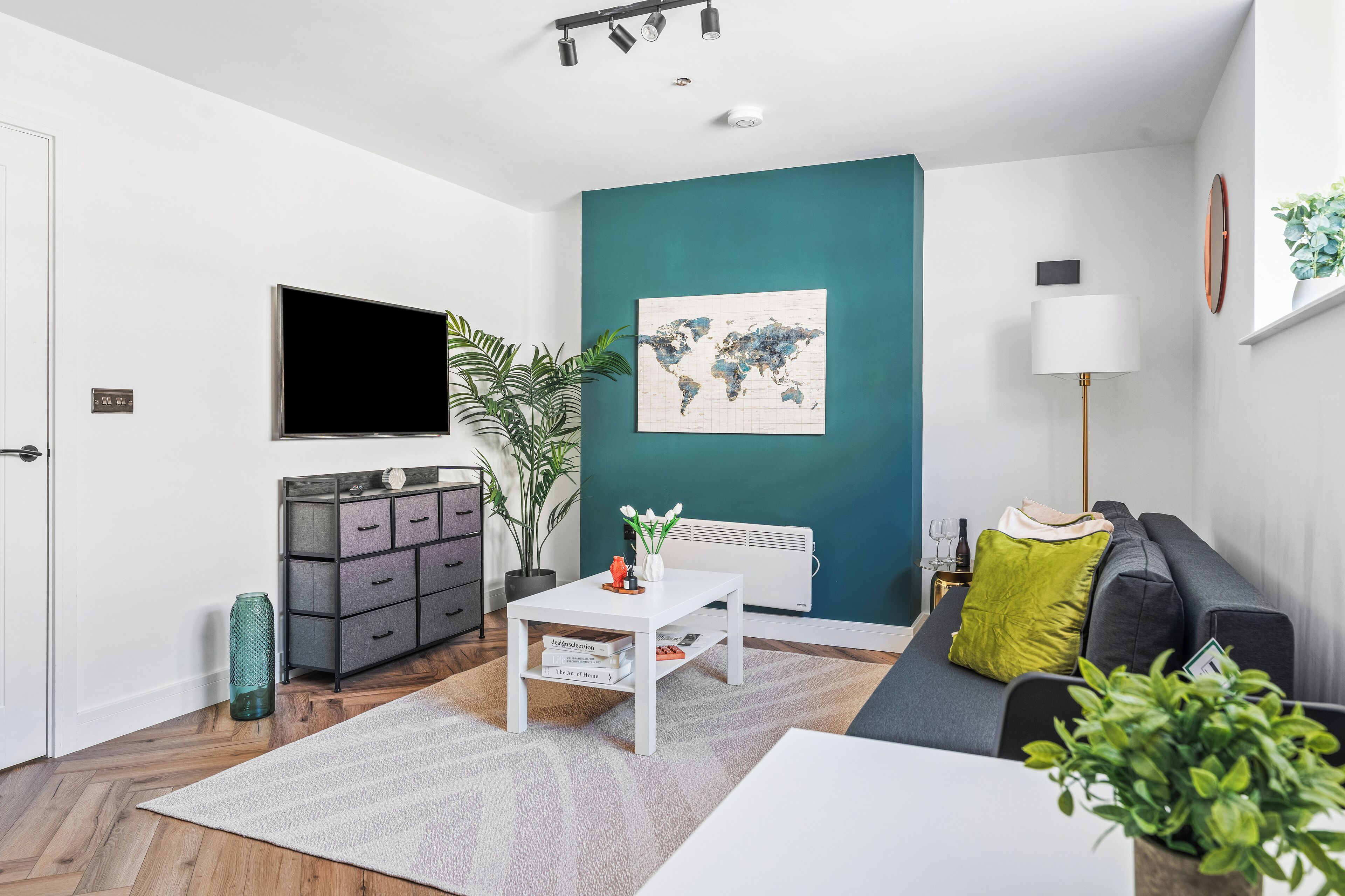 Un salon moderne avec un téléviseur à écran plat, une œuvre d’art représentant une carte du monde, un canapé gris, une table basse blanche et une plante en pot.