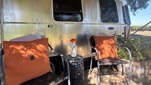 A vintage camper van with two metal chairs and cushions, a small table with a vase, and a window.