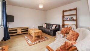 Un salon avec une table basse en bois, un canapé en cuir, un fauteuil blanc, une bibliothèque et un téléviseur à écran plat fixé au mur.