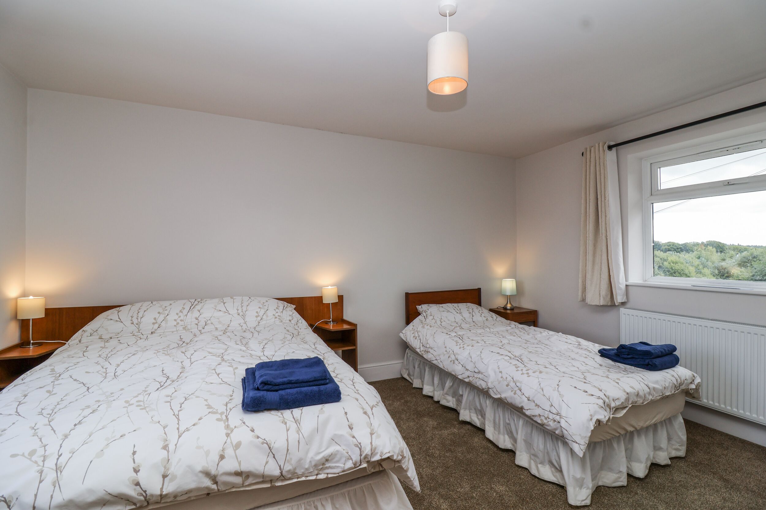 Une chambre d’hôtel avec deux lits simples, chacun doté d’une table de chevet et d’une lampe. La chambre dispose d’une grande fenêtre avec des rideaux, d’un radiateur et d’un luminaire au plafond.
