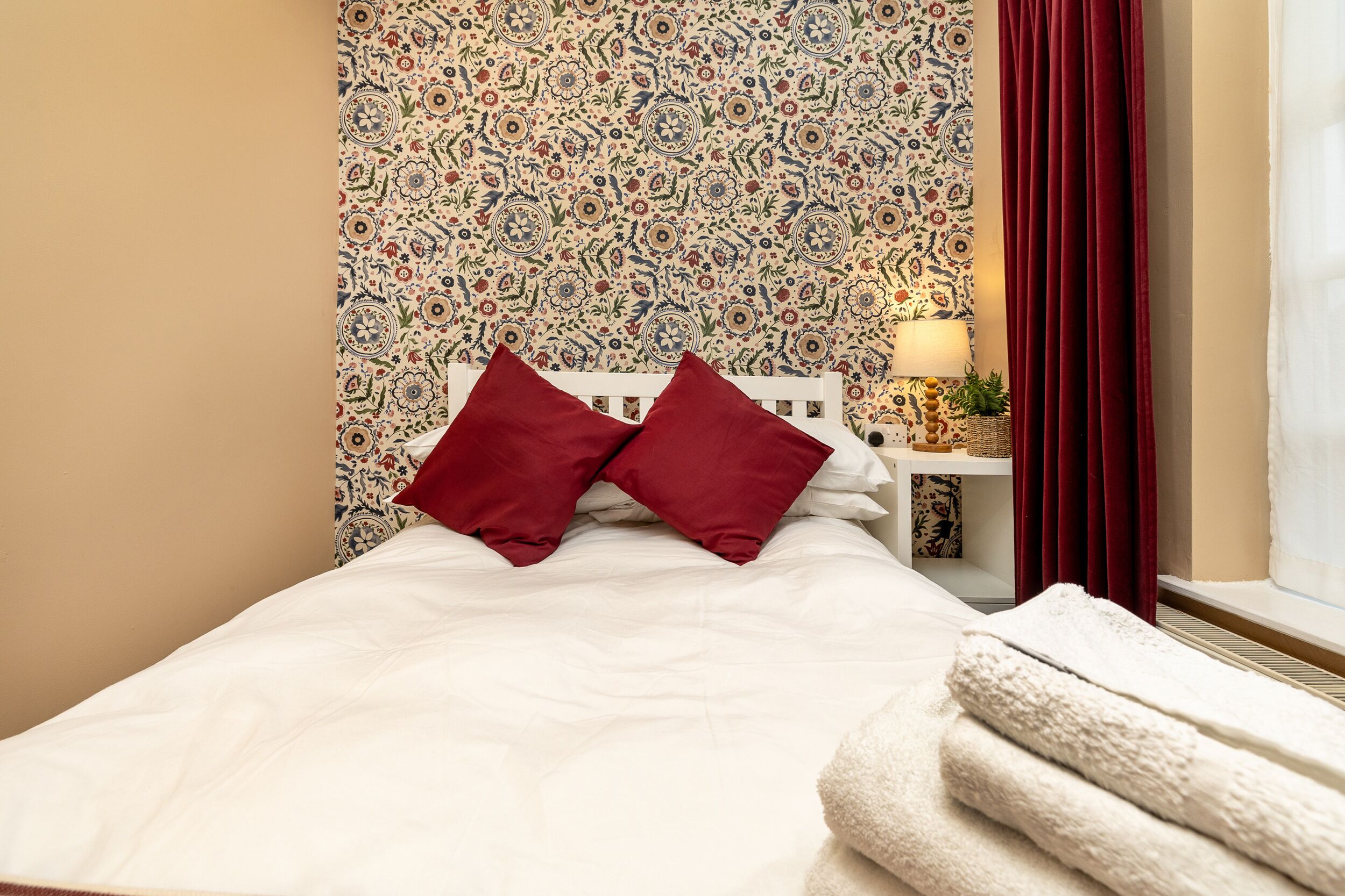 A neatly made bed with white linens and two red pillows, a bedside table with a lamp and a small plant, and a patterned wallpaper.