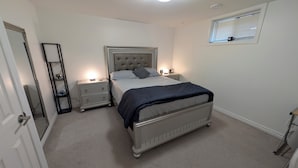 A bedroom with a bed, two bedside tables, a nightstand, a mirror, and a window with blinds. - Spacious one bedroom Basement Apartment (Belleville)