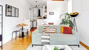 A modern living room with a sofa, coffee table, and a dining area with chairs. A kitchen is visible in the background. - Chic 1BR in the Heart of Le Marais (Paris)
