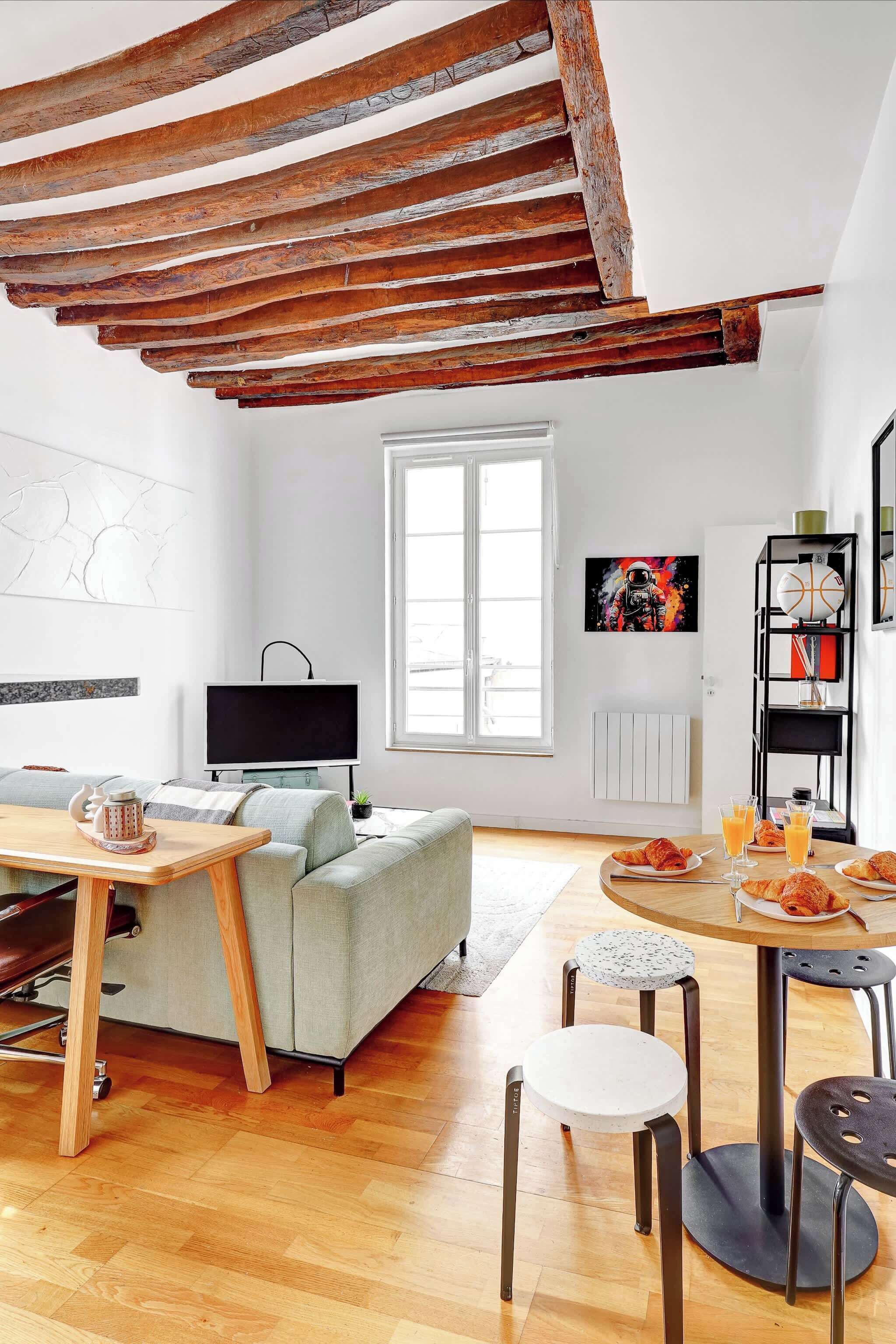Una sala de estar moderna con vigas de madera a la vista, un sofá gris, una mesa de centro de madera, un televisor y un comedor con una mesa redonda y taburetes.