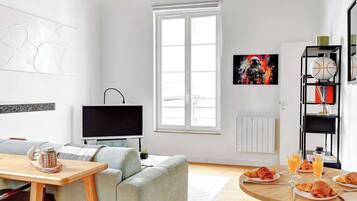 Una sala de estar moderna con vigas de madera a la vista, un sofá gris, una mesa de centro de madera, un televisor y un comedor con una mesa redonda y taburetes.