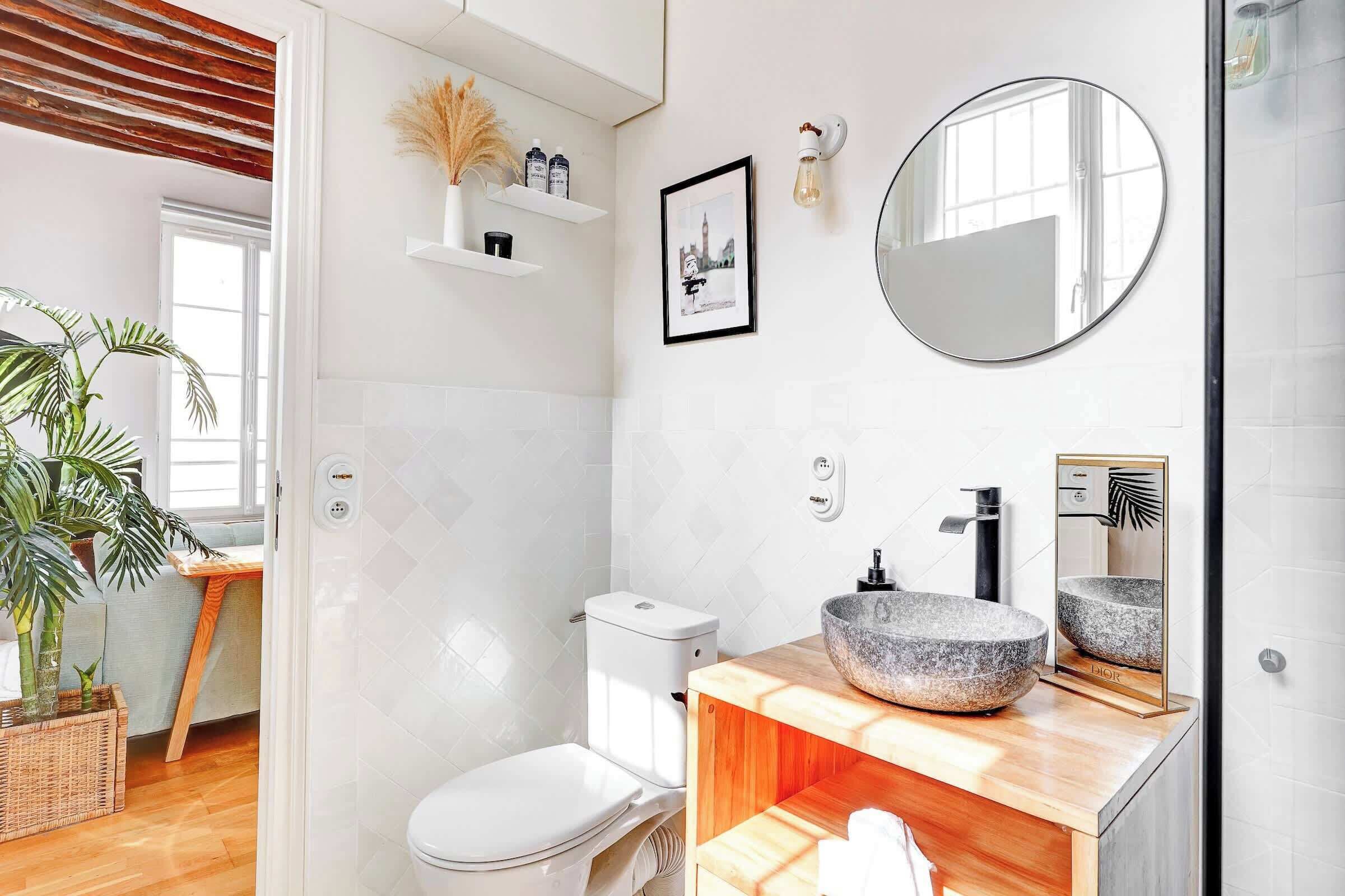 Un baño moderno con inodoro blanco, tocador de madera, espejo redondo y zona de ducha.