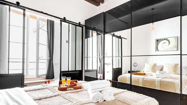 A modern hotel room with a large bed, a wooden bench, a tray with breakfast, and a mirror wall. - Chic 1BR in the Heart of Le Marais (Paris)