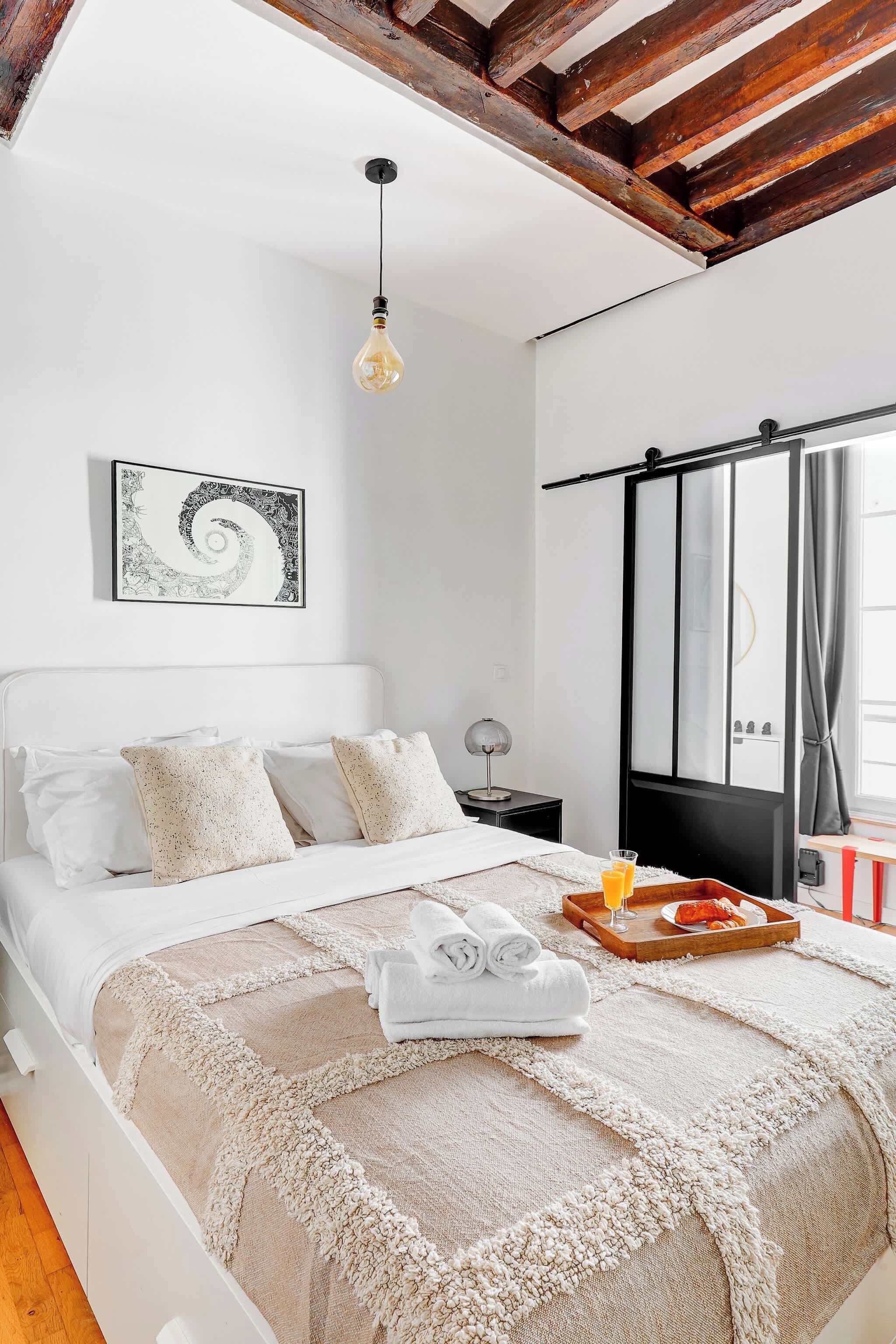 Une chambre à coucher avec un lit, une lampe suspendue, un tableau encadré, une armoire et un plateau avec un verre de jus d’orange.