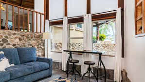 A room with a stone wall, a blue sofa, a patterned rug, a table with two stools, and a window with curtains.
