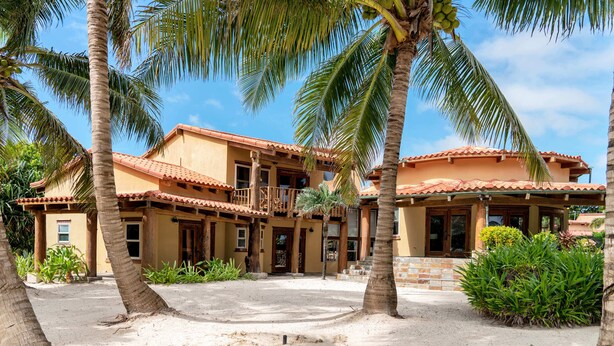 A two-story building with a tiled roof, surrounded by palm trees and a sandy courtyard.
