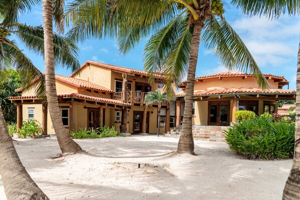 A two-story building with a tiled roof, surrounded by palm trees and a sandy courtyard.