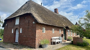 A brick building with a thatched roof, a white door, and a small table with a chair outside. - Holiday Home 'Deichkate' – Vacation in a Historic Monument with Panoramic Views (Dagebüll)