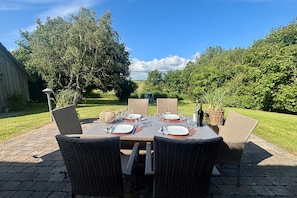 Un área de comedor al aire libre con sillas y mesas de mimbre, rodeada de vegetación y un cielo despejado.