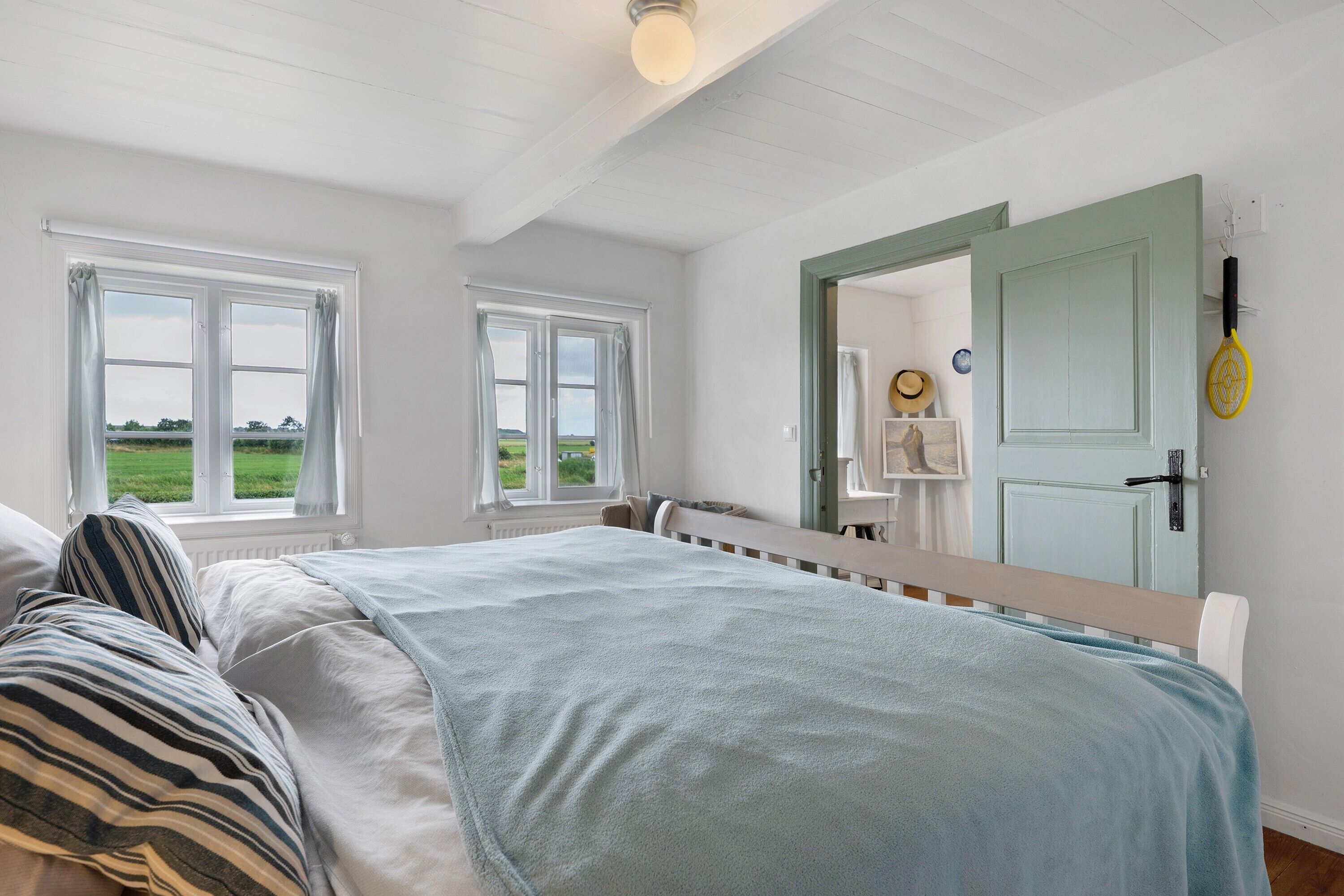 A bedroom with a bed, a window, a door, and a tennis racket on the wall.