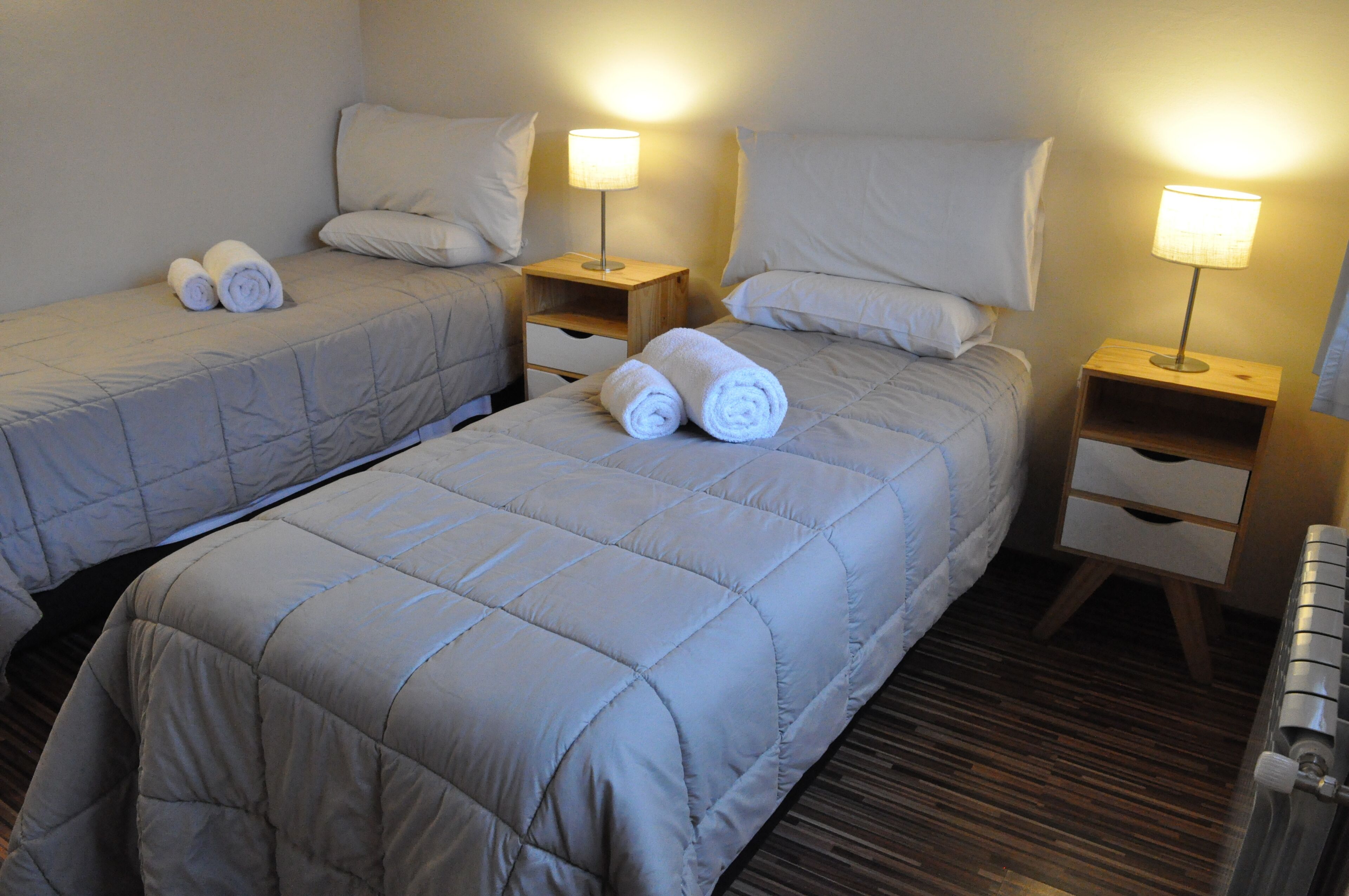 Habitación de hotel con dos camas individuales, cada una con una almohada, una mesita de noche con lámpara y toallas enrolladas sobre las camas.