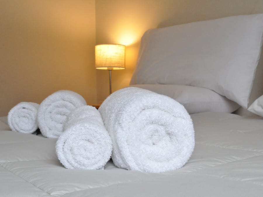 A neatly made bed with rolled white towels, a lamp, and a plain wall.
