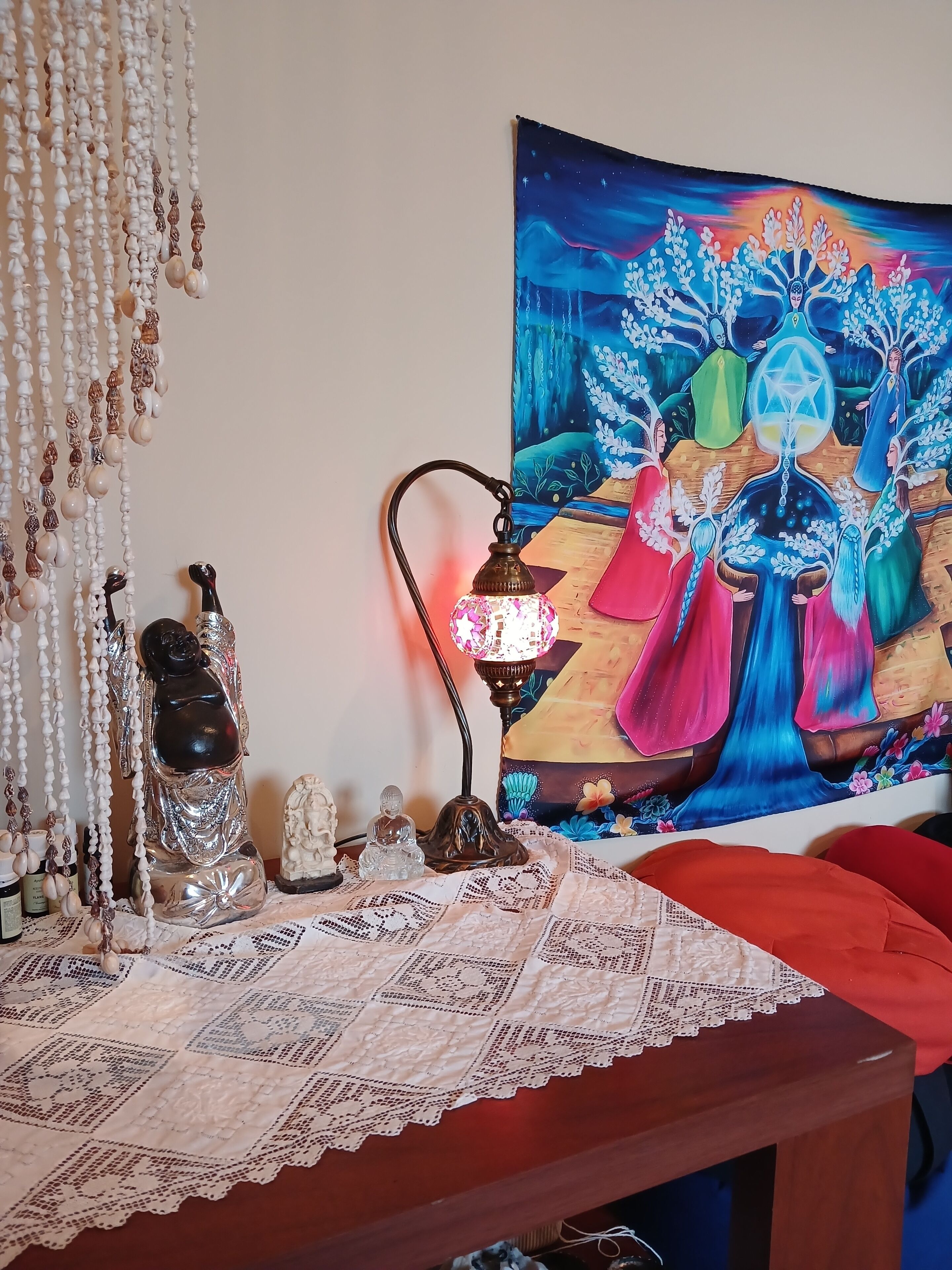 A room with a table decorated with a lace tablecloth, a Buddha statue, a lamp, and a tapestry depicting a colorful scene.
