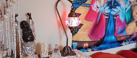 A room with a table decorated with a lace tablecloth, a Buddha statue, a lamp, and a tapestry depicting a colorful scene.