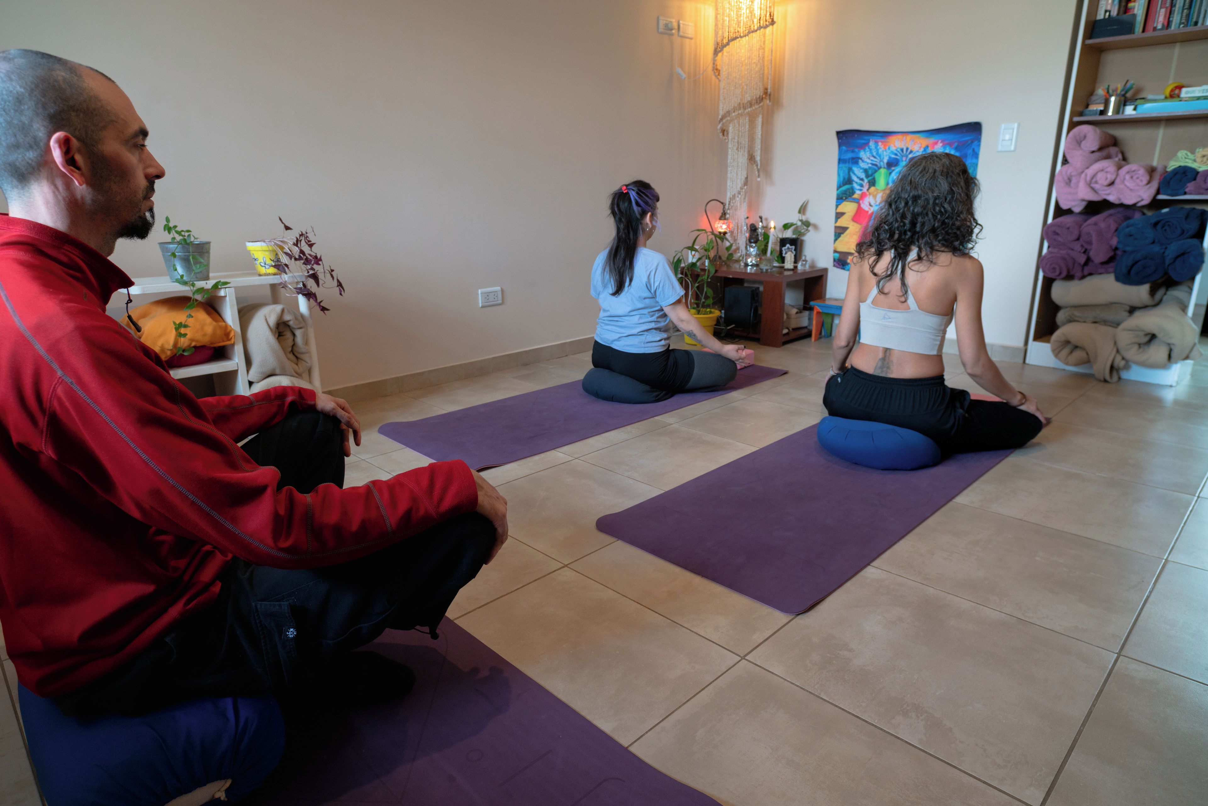 Three individuals seated on purple exercise mats in a room with a tiled floor, a bookshelf, and a wall with a tapestry.